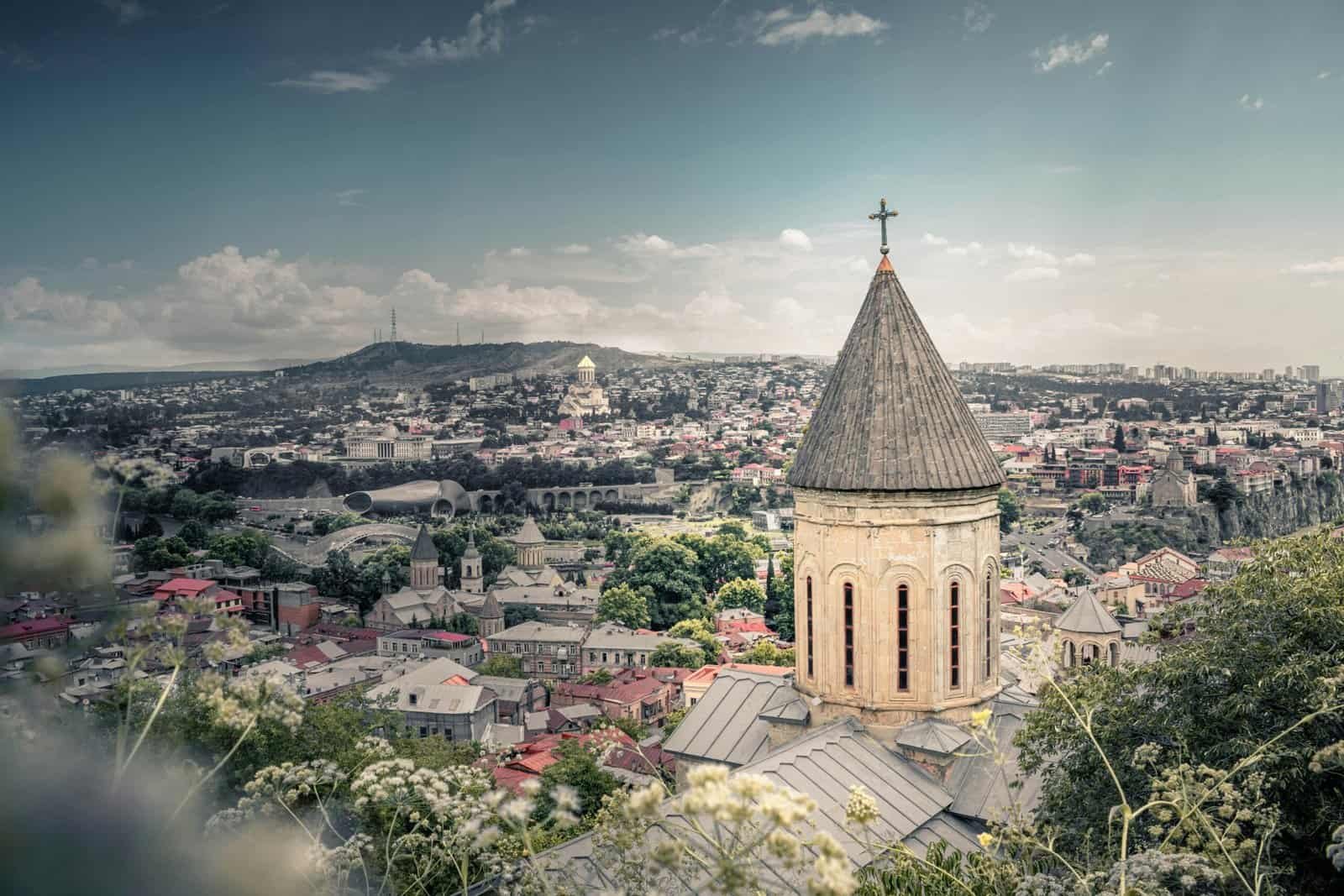 View of a historical church tower with Tbilisi's cityscape in the background under a blue sky.